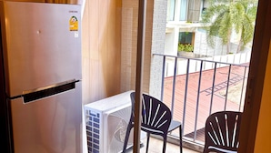 Une pièce équipée d’un réfrigérateur Samsung, d’un climatiseur mobile blanc, de deux chaises noires, d’un tapis beige et d’un balcon offrant une vue sur un immeuble et des palmiers.