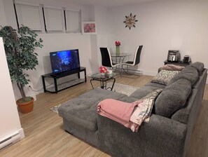A living room with a grey sofa, a TV, a potted plant, a coffee machine, and a dining area with chairs and a table. - Charming Haven House (Moncton)