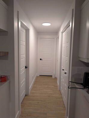 A narrow hallway with white doors, a wooden floor, and a ceiling light. - Charming Haven House (Moncton)