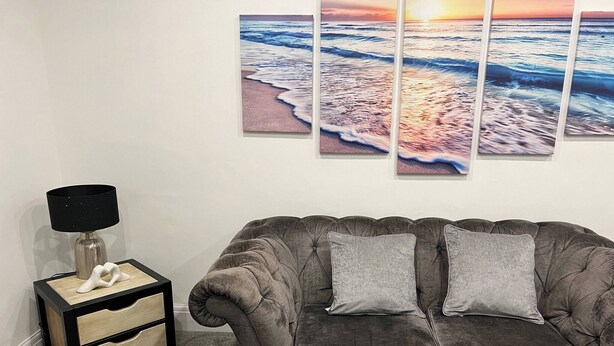 A living room with a gray sofa, two pillows, a small side table with a lamp, and three framed pictures on the wall. - Salvation View 2 Bed Apartment (Hadleigh)