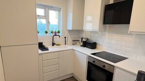 A compact kitchen with white cabinets, a black stove, and a window with plants. - Salvation View 2 Bed Apartment (Hadleigh)