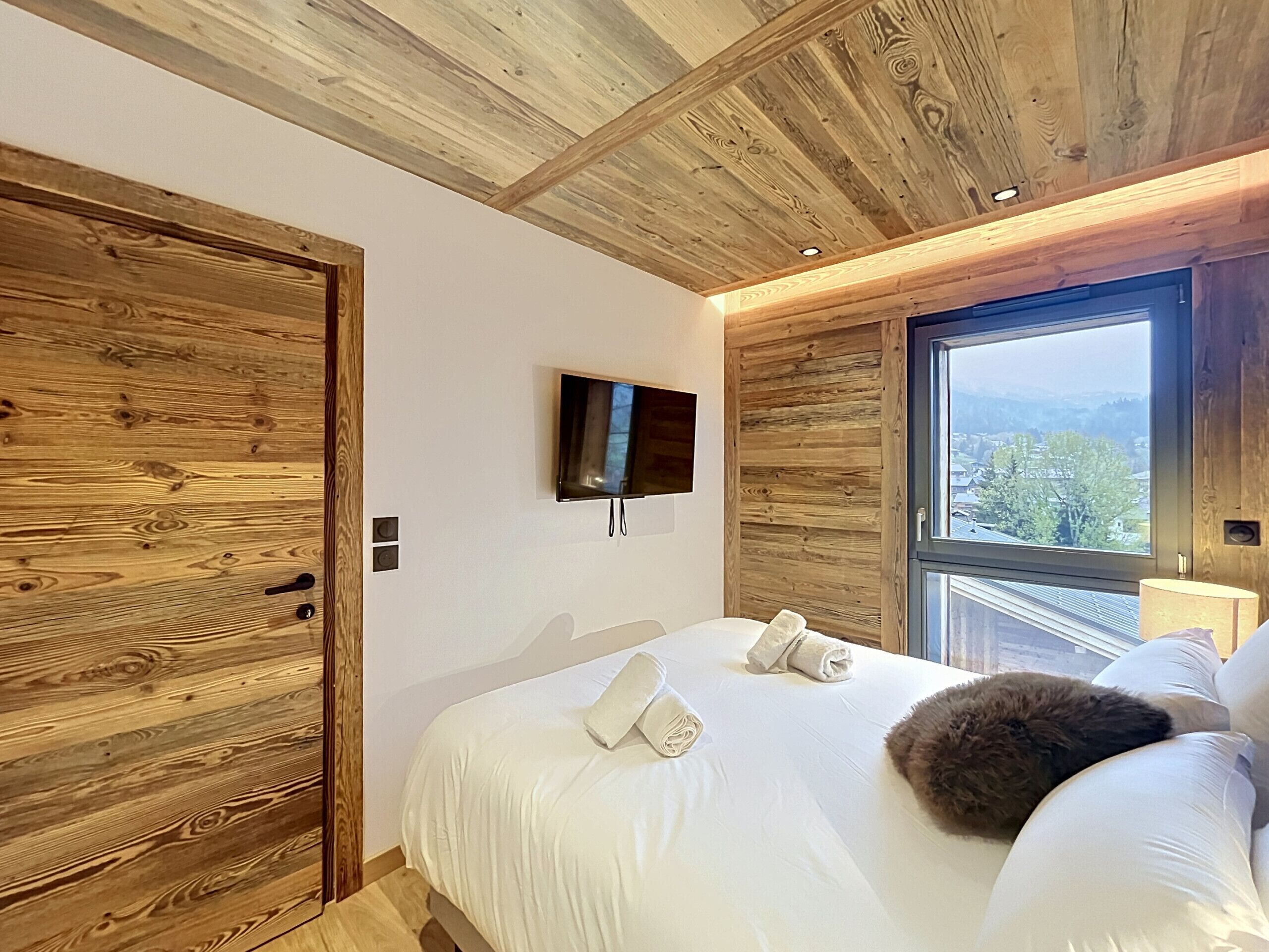 A bedroom with a wooden ceiling, a bed with white linens, a TV mounted on the wall, and a window with a view of the outside.