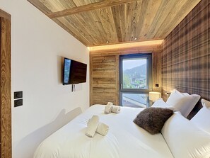A bedroom with a wooden ceiling, a television mounted on the wall, a large window with a view of buildings and hills, and a bed with white linens and pillows. - The Douka Apartment—Sleeps 4—Near Downtown (Les Gets)
