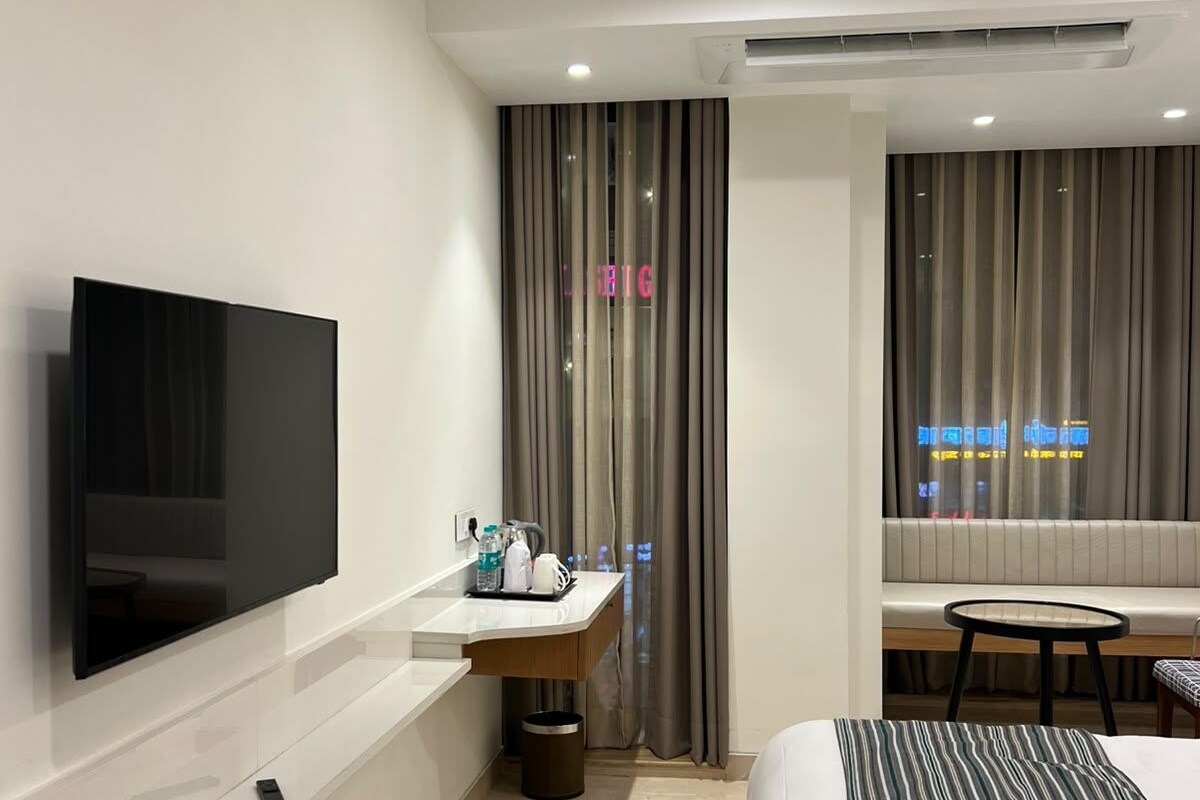 A modern hotel room with a flat-screen TV mounted on the wall, a bedside table with a lamp, a small round table, and a bed with striped bedding.