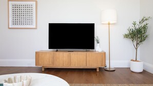 Ein Wohnzimmer mit Flachbildfernseher auf einer Holzkabine, einer Stehlampe, einem Blumentopf und einem gerahmten Bild an der Wand.