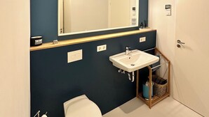 A modern bathroom with a dark blue wall, a white sink, a toilet, and a mirror with lights.