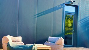 A modern outdoor seating area with two sofas and a wooden table, set against a corrugated metal wall with a door and window.