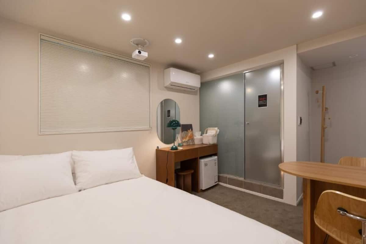 A hotel room with a bed, a desk with a chair, a mirror, a small refrigerator, and a sliding door closet.