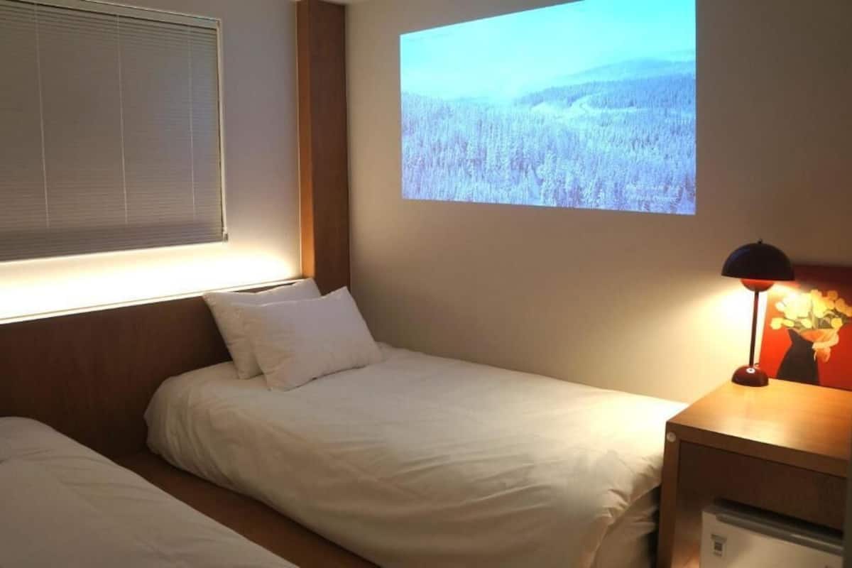 A hotel room with two beds, a projector displaying a forest landscape, a nightstand with a lamp, and a window with blinds.