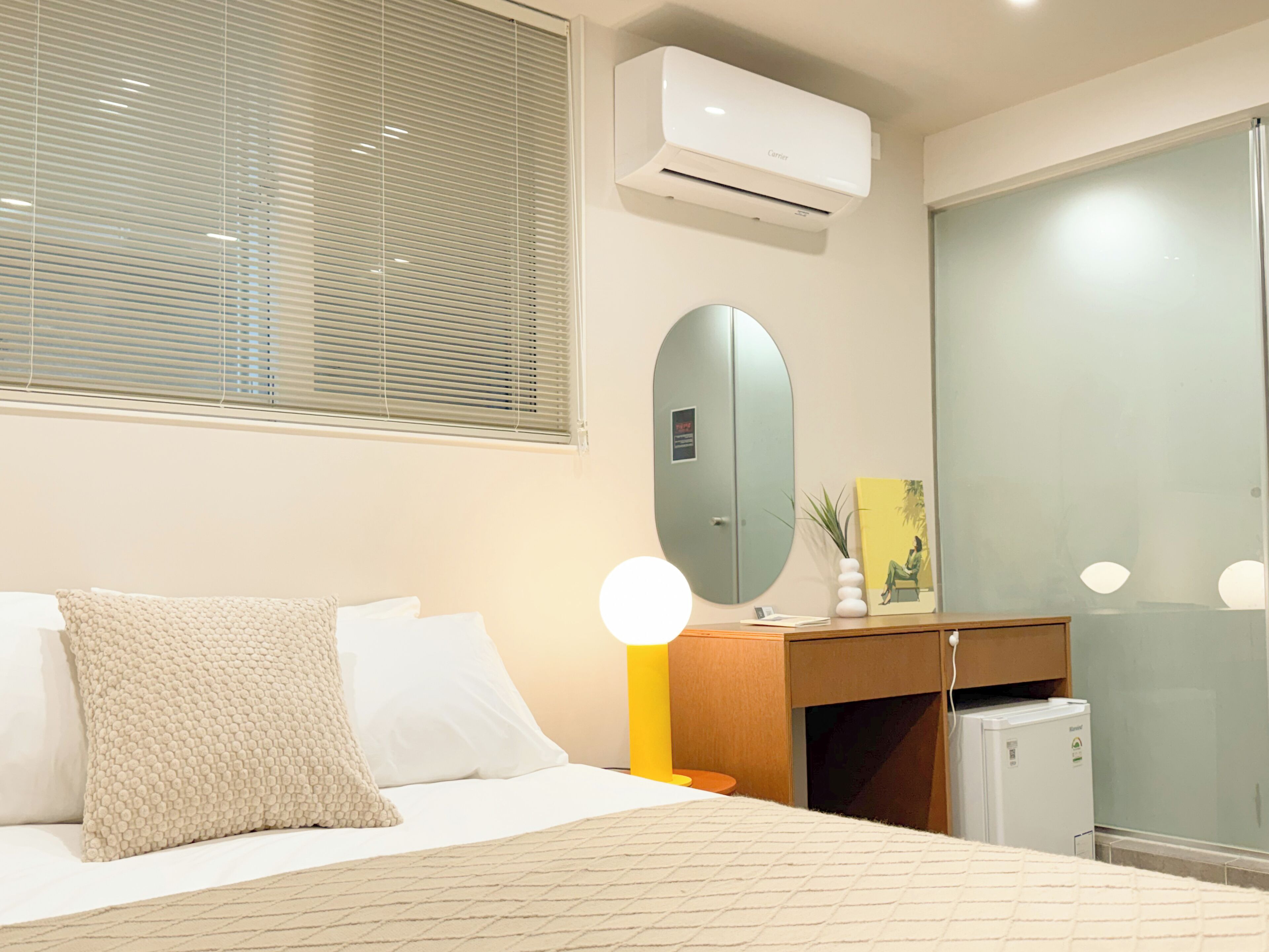 A hotel room with a bed, bedside table, lamp, mirror, and a small refrigerator.