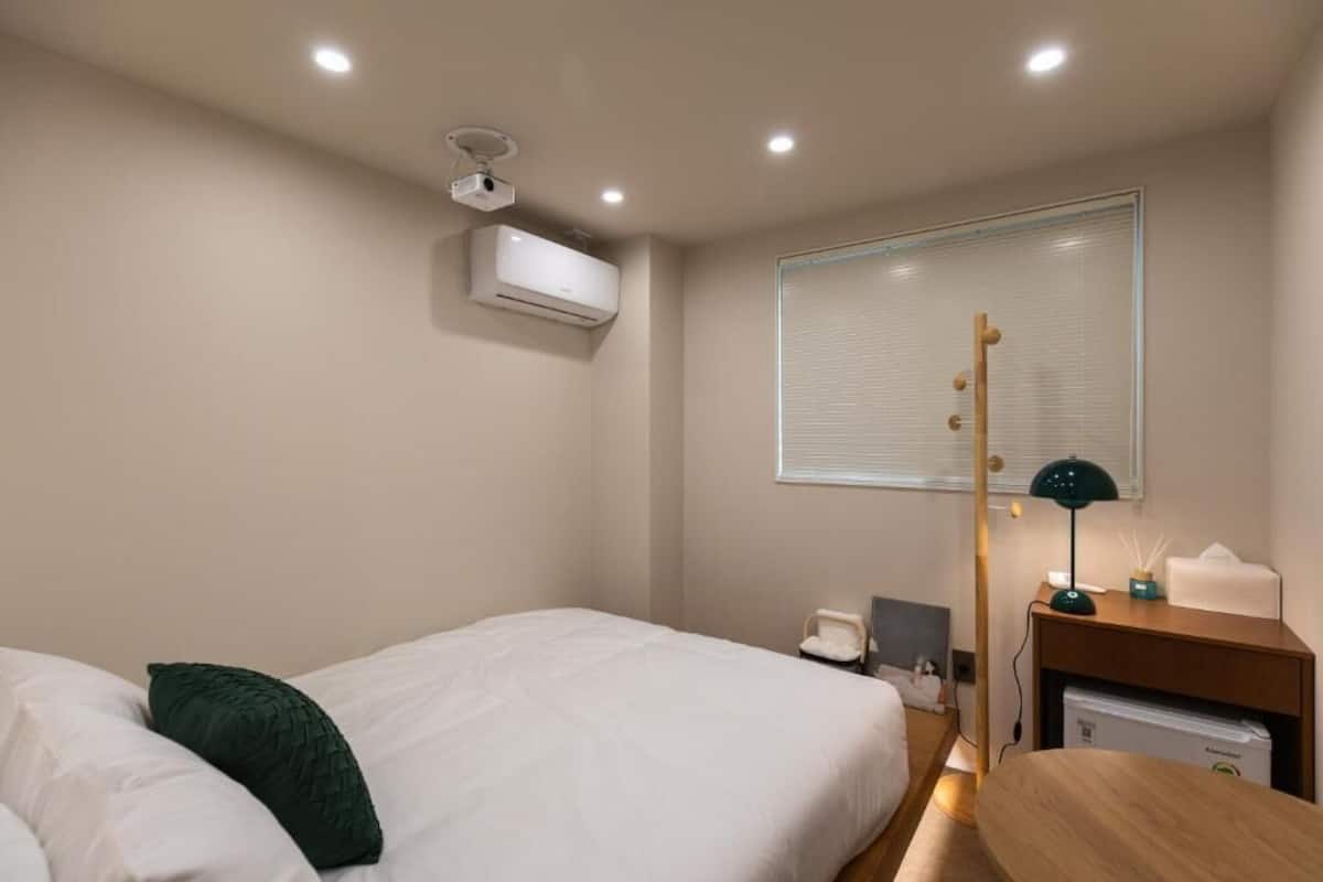 A hotel room with a bed, a desk, a lamp, a small table, an air conditioning unit, a window with blinds, and a ceiling projector.