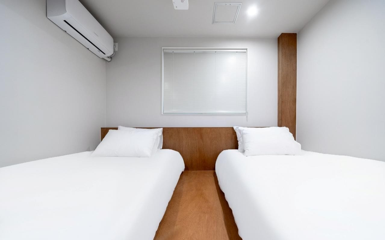 A hotel room with two beds, a wooden headboard, a window with blinds, and an air conditioning unit.