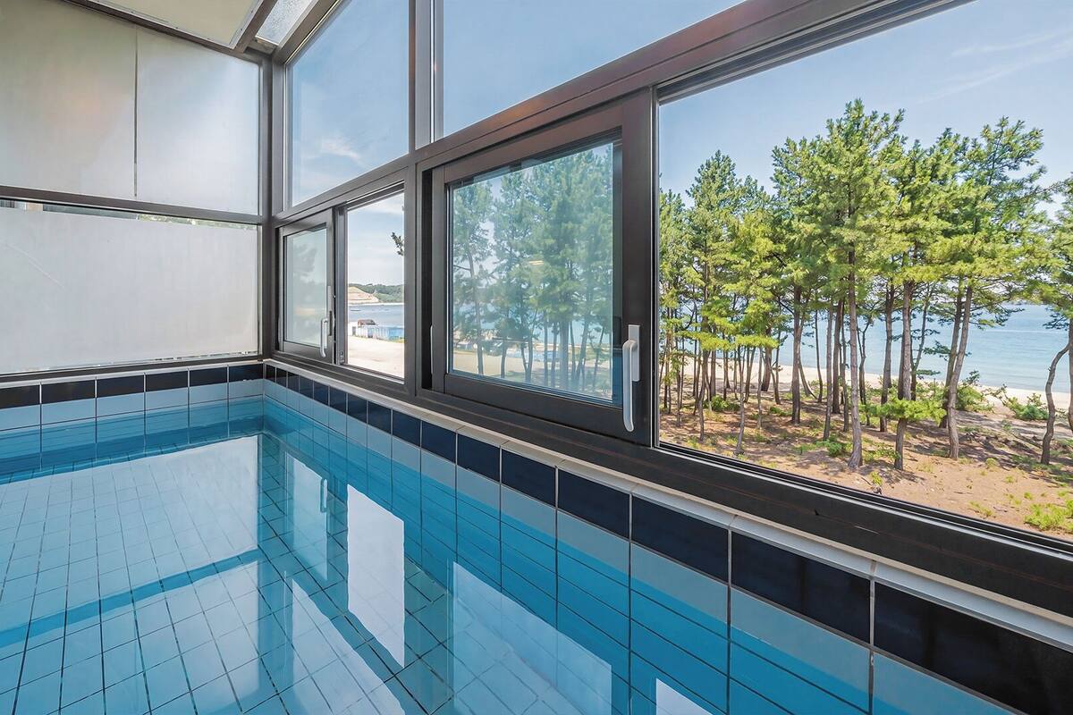 Una piscina con baldosas azules brillantes, ventanas amplias con marcos negros y vistas a una playa con árboles y el mar.