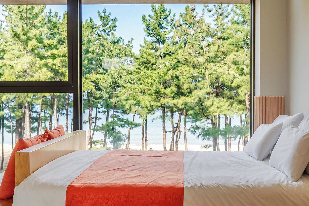 Un dormitorio con un ventanal grande, una cama con ropa de cama blanca y naranja, y vistas a árboles y agua.