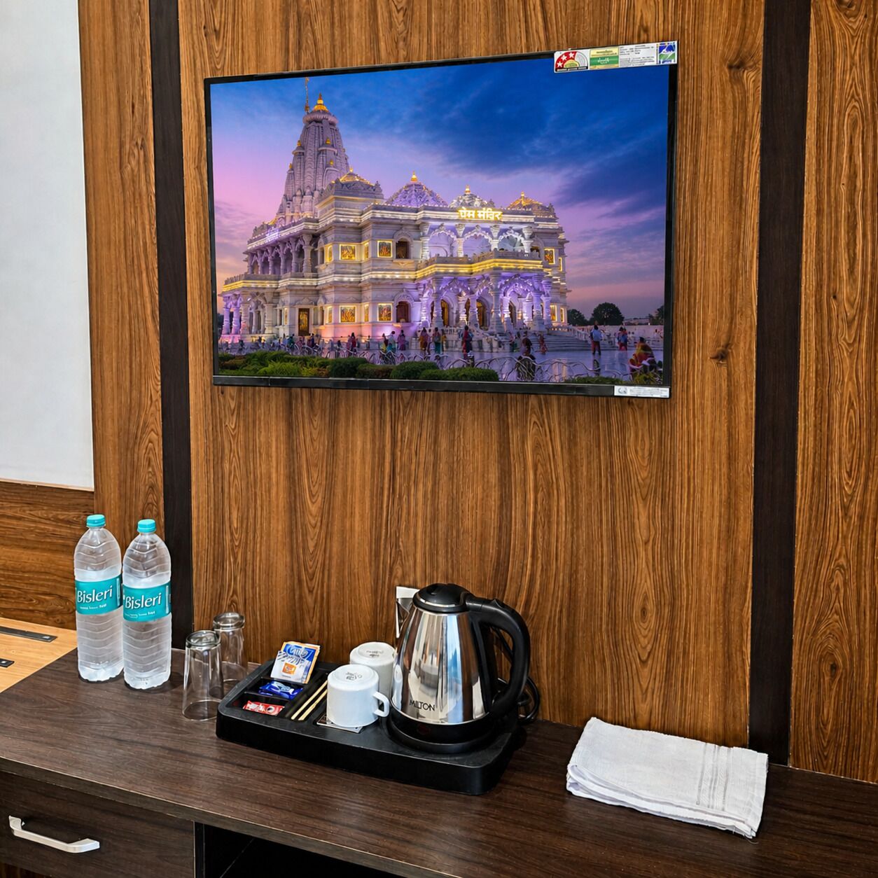 Habitación de hotel con pared de paneles de madera, un televisor de pantalla plana mostrando un templo, una bandeja con una tetera, botellas de agua Bisleri y una toalla doblada.