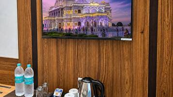 Habitación de hotel con pared de paneles de madera, un televisor de pantalla plana mostrando un templo, una bandeja con una tetera, botellas de agua Bisleri y una toalla doblada.