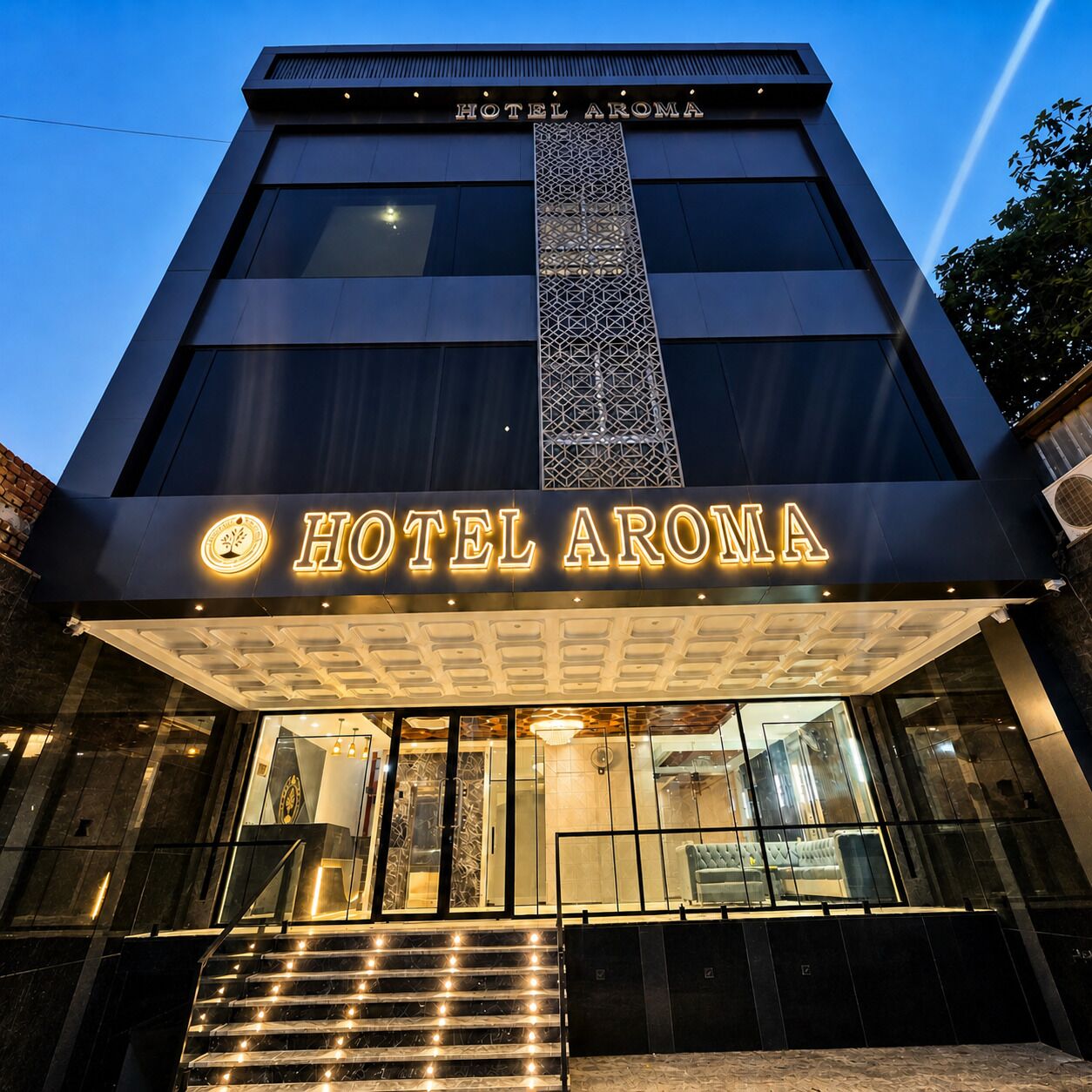A modern hotel building with a prominent 'HOTEL AROMA' sign, large windows, and a central decorative column.