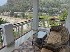 A balcony with a wicker sofa and a glass table, overlooking a hilly landscape with buildings.