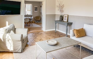 A living room with a sofa, a coffee table, a dining area with chairs, and a kitchen visible through an open door.