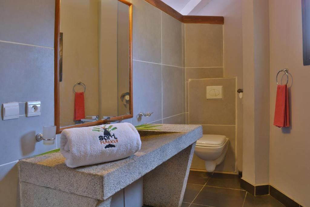Une salle de bain avec un comptoir en pierre, des toilettes blanches, un miroir et une serviette ornée d’un logo.