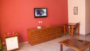 A room with a television mounted on the wall, a wooden cabinet, a small refrigerator, a wooden table, and a chair. - Hotel Residence Sarimanok (Nosy Be)