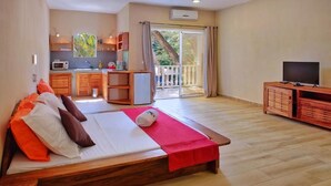 A bedroom with a wooden bed, a TV, a balcony, and a kitchenette. - Hotel Residence Sarimanok (Nosy Be)