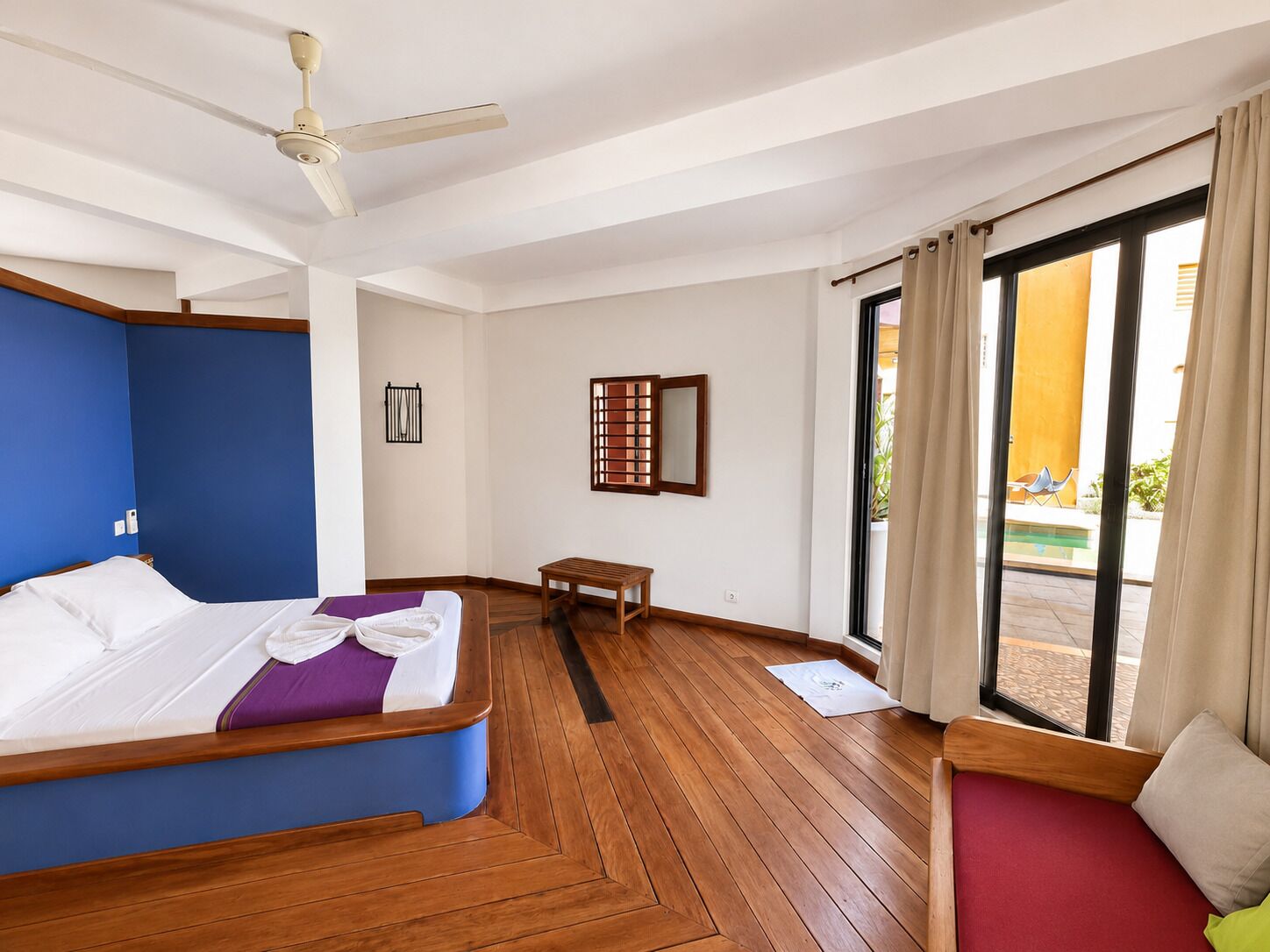 Habitación de hotel con cama, ventilador de techo, ventana con cortinas y vista al exterior.