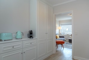 A hallway with a kitchenette area featuring a toaster, kettle, and coffee maker. There is a door leading to a living area with a sofa and a lamp. - Beach Suite - Holiday apartment "Beach Suite" in Westerland (Sylt)