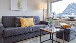 Ein modernes Wohnzimmer mit einem grauen gesteppten Sofa, einem gelben Kissen, einem Glastisch und Blick auf eine Stadtansicht durch ein Fenster.