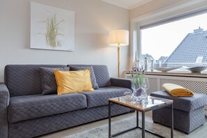 A modern living room with a grey quilted sofa, a yellow pillow, a glass coffee table, and a view of a cityscape through a window. - Beach Suite - Holiday apartment "Beach Suite" in Westerland (Sylt)