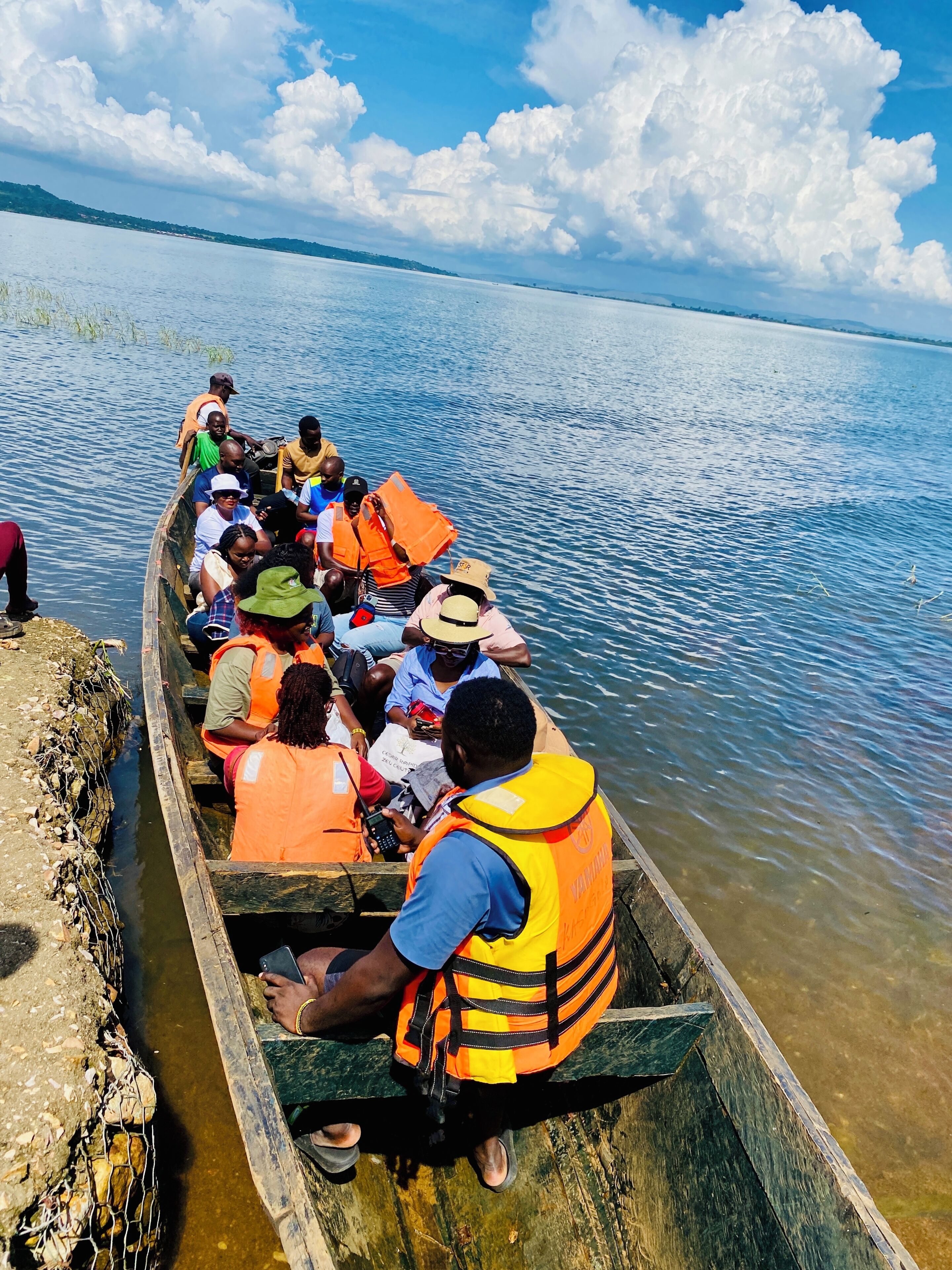 Un grupo de personas en un bote de madera con chalecos salvavidas, con un cuerpo de agua tranquilo y un cielo despejado al fondo.