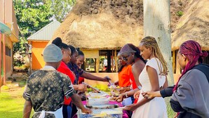 Un grupo de personas se sirve de una mesa de buffet al aire libre, con una estructura de techo de paja al fondo.