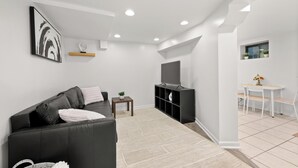 Un salon moderne avec un canapé noir, un téléviseur, une petite table ornée d’une plante, et un coin repas comprenant une table et des chaises.