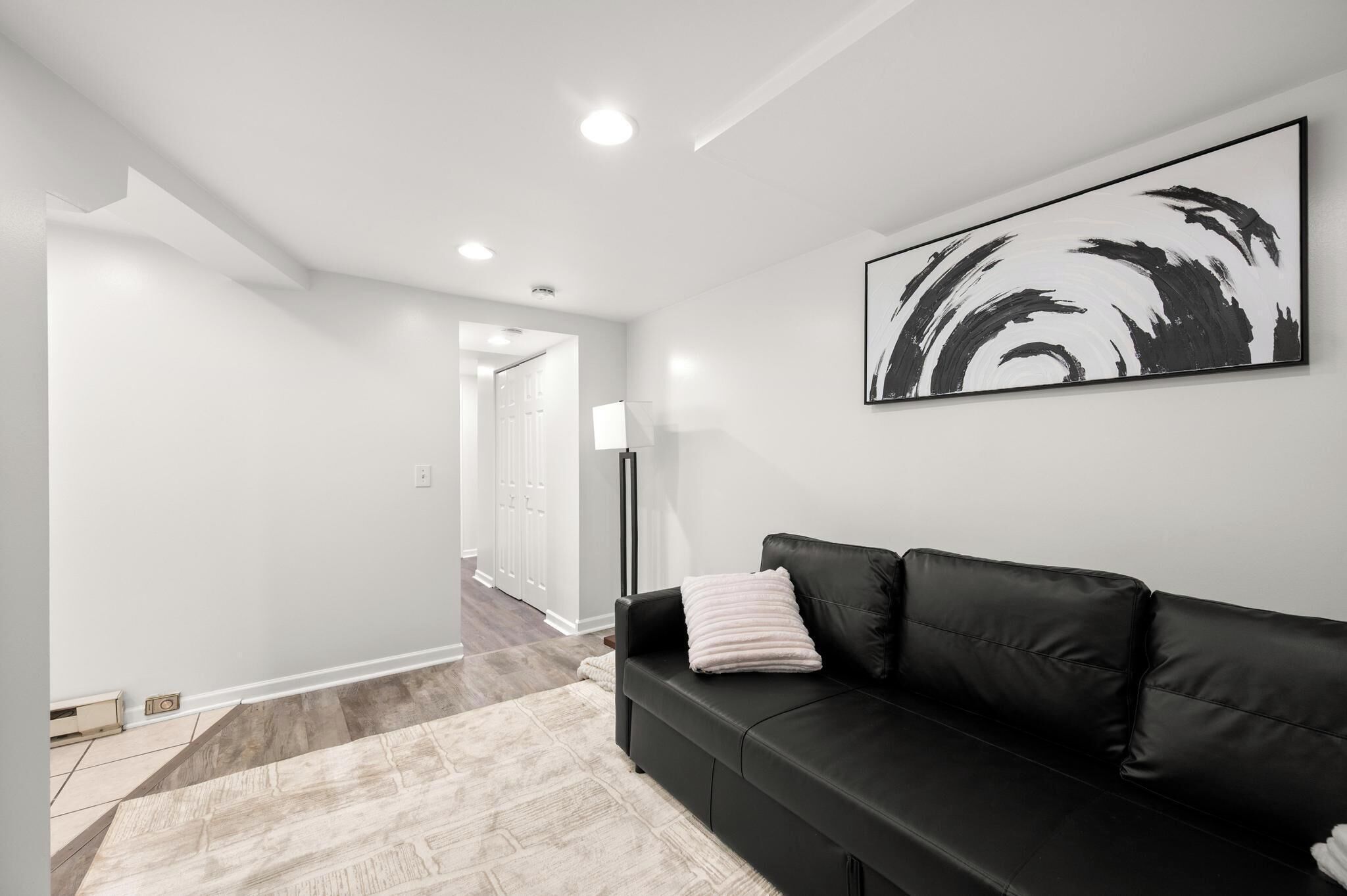 A modern living room with a black leather sofa, a large abstract painting, and a hallway leading to another room.