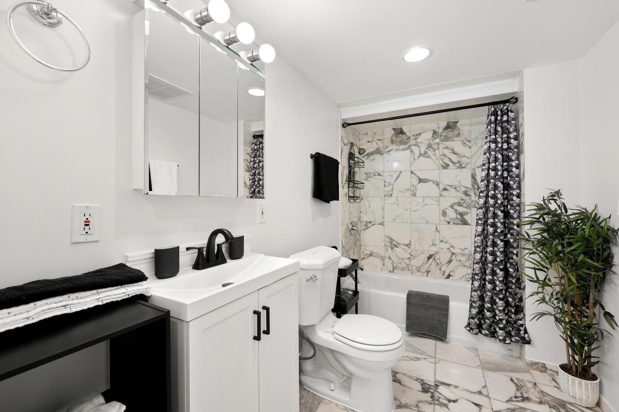 A modern bathroom with a white sink, black faucet, and a mirror with lights. There's a toilet, a bathtub with a shower curtain, and a potted plant.