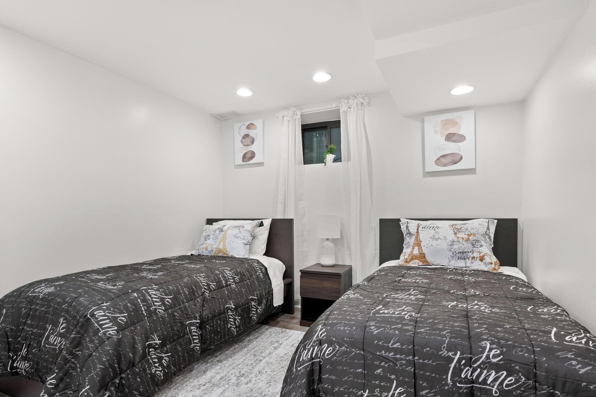 A bedroom with two beds, white walls, a window with curtains, and artwork on the walls.