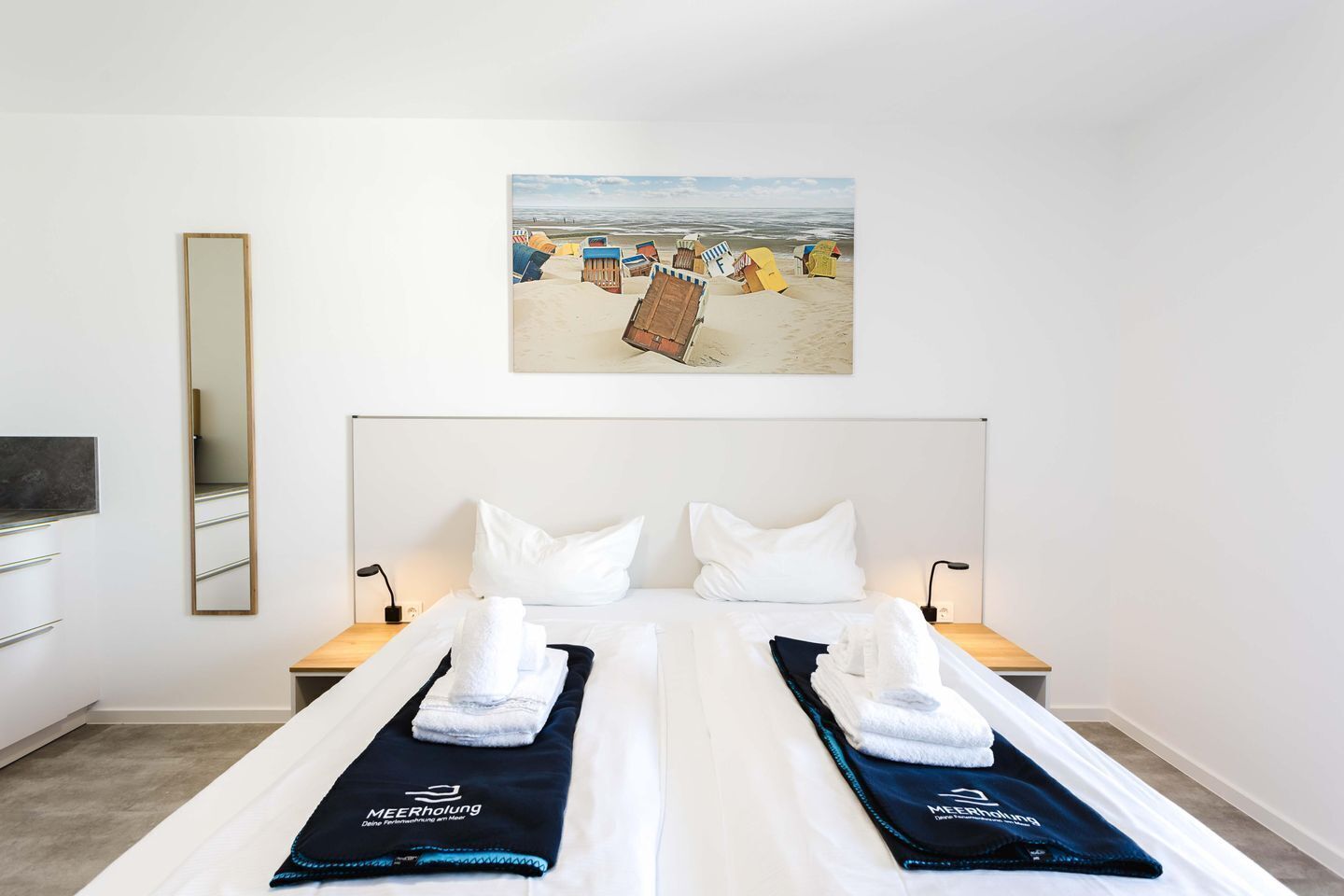 Ein Hotelzimmer mit einem Bett, zwei Nachttischen, einem Spiegel und einem Wandbild mit Strandmotiv.