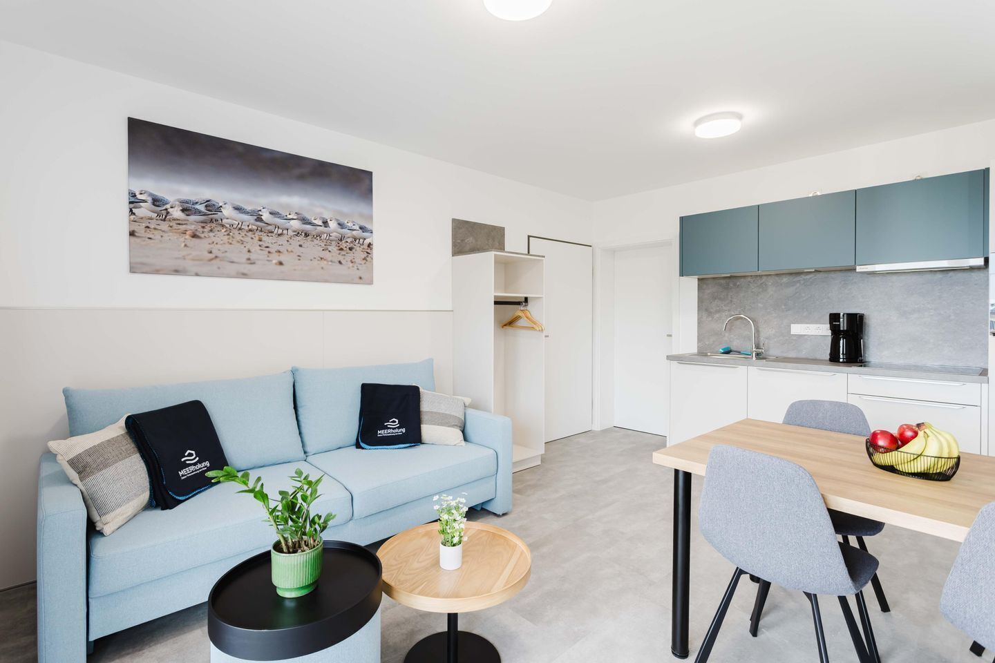 Ein modernes Wohnzimmer mit Sofa, Couchtisch und Essbereich mit Stühlen und Tisch. Es gibt eine Küche mit Schränken und Spüle.