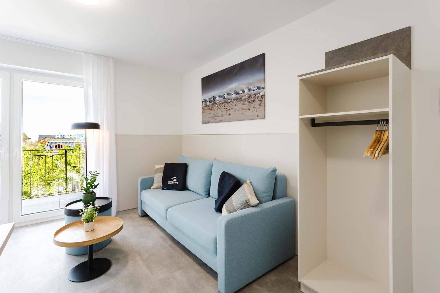 Una sala moderna con un sofá azul claro, una mesa de centro redonda de madera, una planta en maceta, un cuadro en la pared y vistas a través de una ventana.