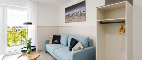 Una sala moderna con un sofá azul claro, una mesa de centro redonda de madera, una planta en maceta, un cuadro en la pared y vistas a través de una ventana.