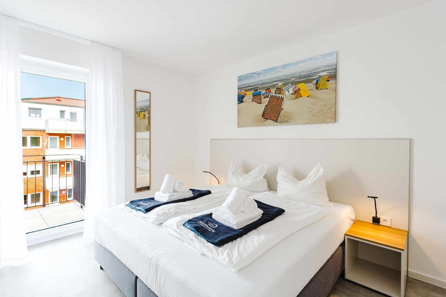 Een hotelkamer met een bed, een schilderij van mensen op het strand, een raam met uitzicht op gebouwen en een nachtkastje met een lamp.