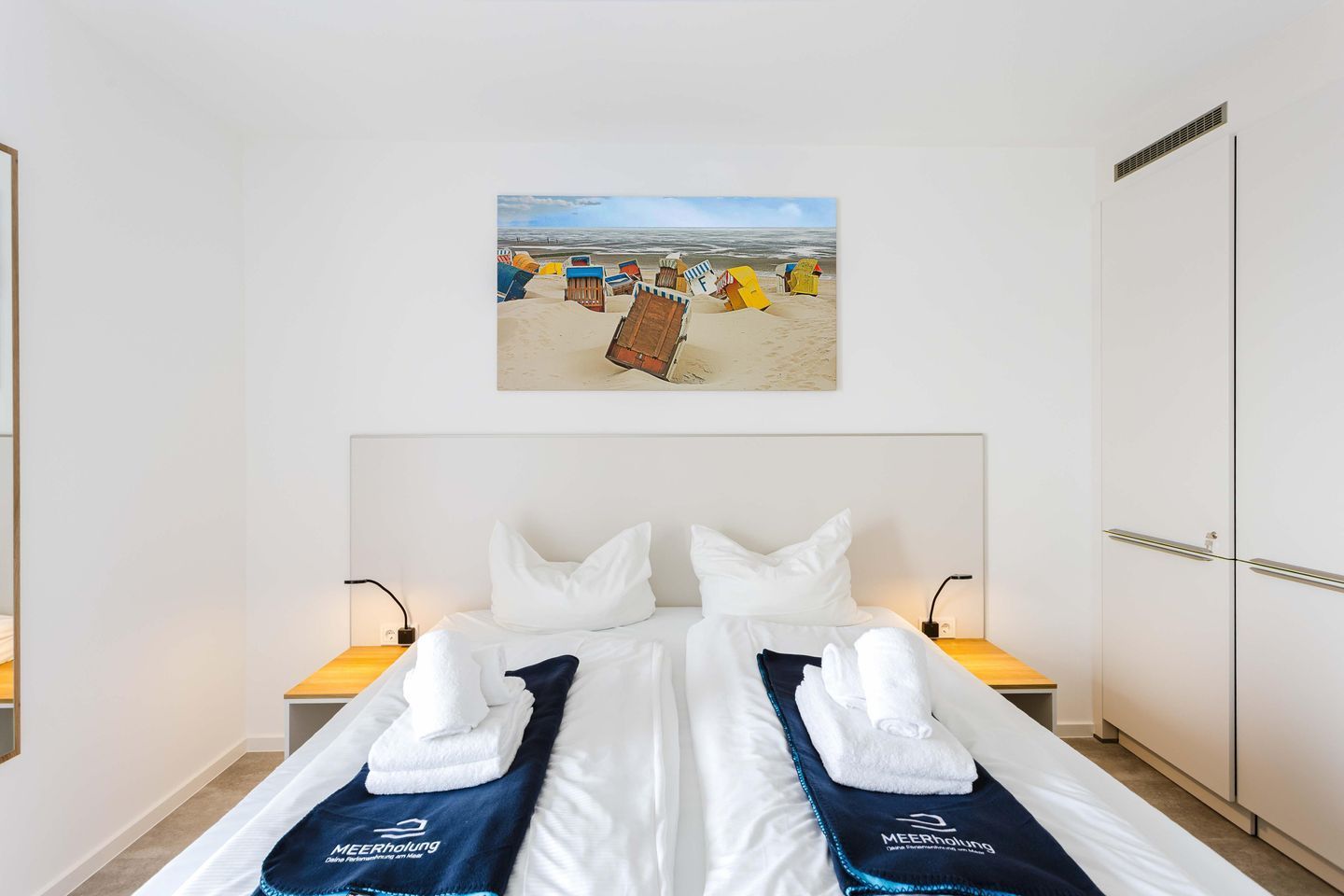 Ein Hotelzimmer mit einem Bett, zwei Nachttischen, einem Wandbild mit Strandmotiv und einem Kleiderschrank.