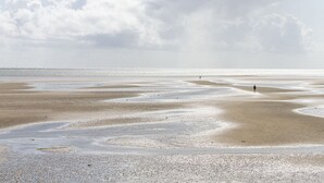 A beach with pebbles and sand, a person walking, and a cloudy sky. - MEERholung Vacation Rental 2.8 with Balcony, Just 400 m from the Sea - MEERholung House (Cuxhaven)