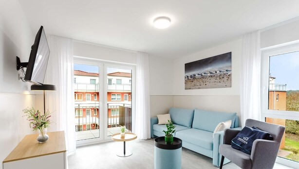 A modern living room with a sofa, armchair, TV, and a view of buildings through large windows. - MEERholung Vacation Rental 2.8 with Balcony, Just 400 m from the Sea - MEERholung House (Cuxhaven)