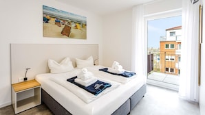 A hotel room with a large bed, a nightstand, a painting of beach huts, and a balcony with a city view. - MEERholung Vacation Rental 2.8 with Balcony, Just 400 m from the Sea - MEERholung House (Cuxhaven)