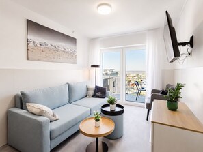 Ein modernes Wohnzimmer mit einer Couch, einem kleinen runden Tisch mit Pflanze, einem an der Wand montierten Fernseher und einem Balkon mit Blick.