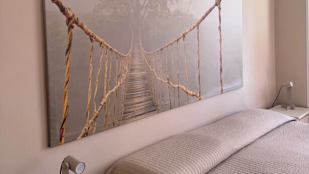 A bedroom with a large painting of a rope bridge, a bed with a beige quilt, and a bedside table with a lamp. - 1.5 km from the AOI airport, comfortable studio apartments for your needs. (Falconara Marittima)