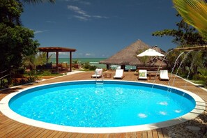 A circular pool with a wooden deck, lounge chairs, and a thatched-roof structure. - Pitinga Hotel Pousada (Guesthouse) (Arraial D'Ajuda - Porto Seguro)