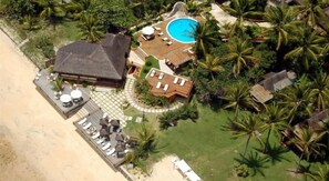 An aerial view of a resort with a swimming pool, a large building, and a beach area with lounge chairs. - Pitinga Hotel Pousada (Guesthouse) (Arraial D'Ajuda - Porto Seguro)