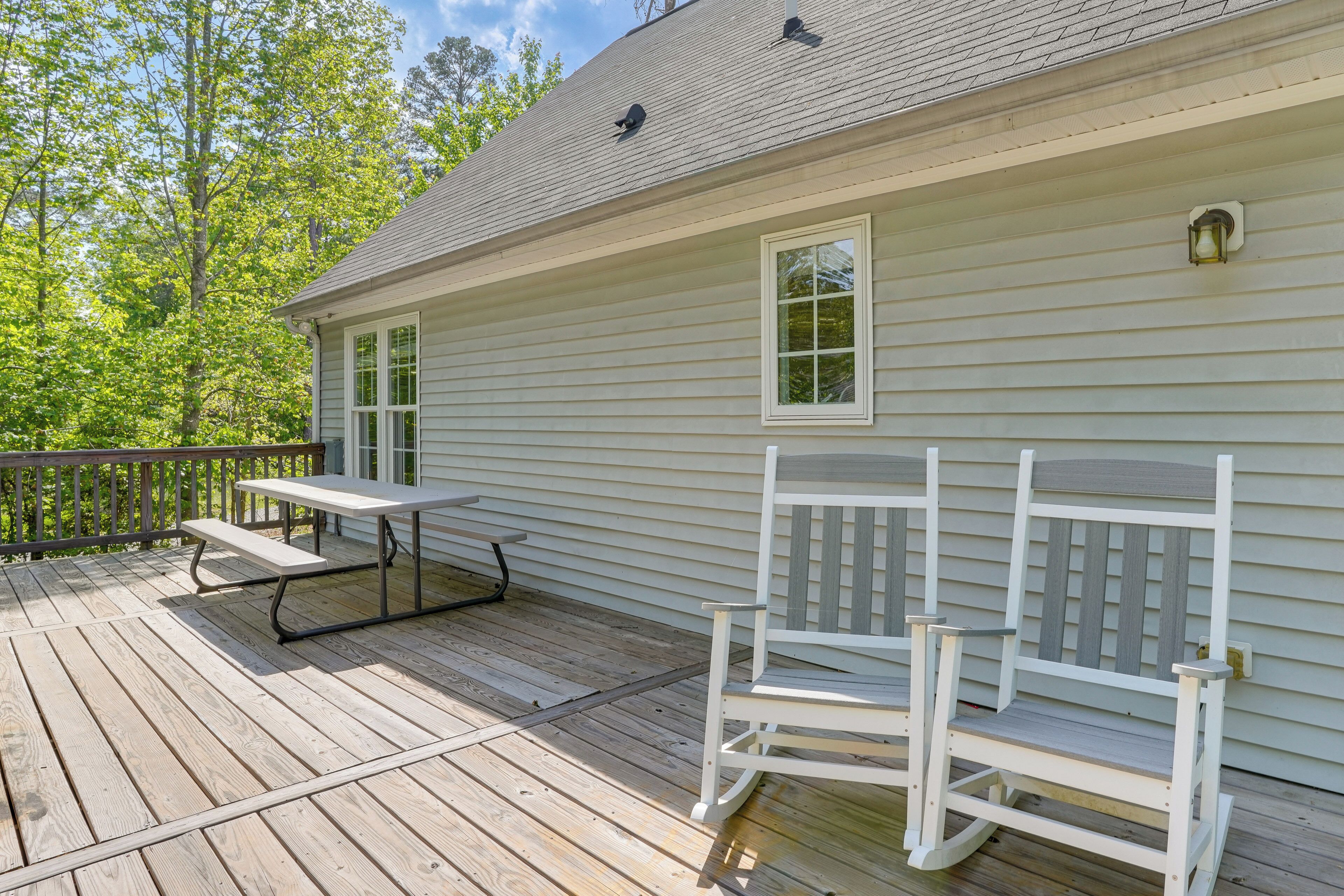En træterrasse med et picnicbord og to Adirondack-stole, et hvidt hus med et gråt tag og et træhegn.
